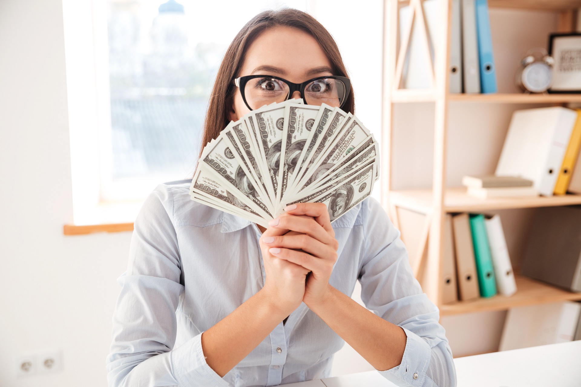 Picture of businesswoman dressed in white shirt sitting in her office and holding money in hand near face